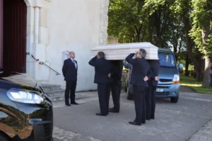 Porteurs de pompes funèbres transportant un cercueil en bois clair devant une église.